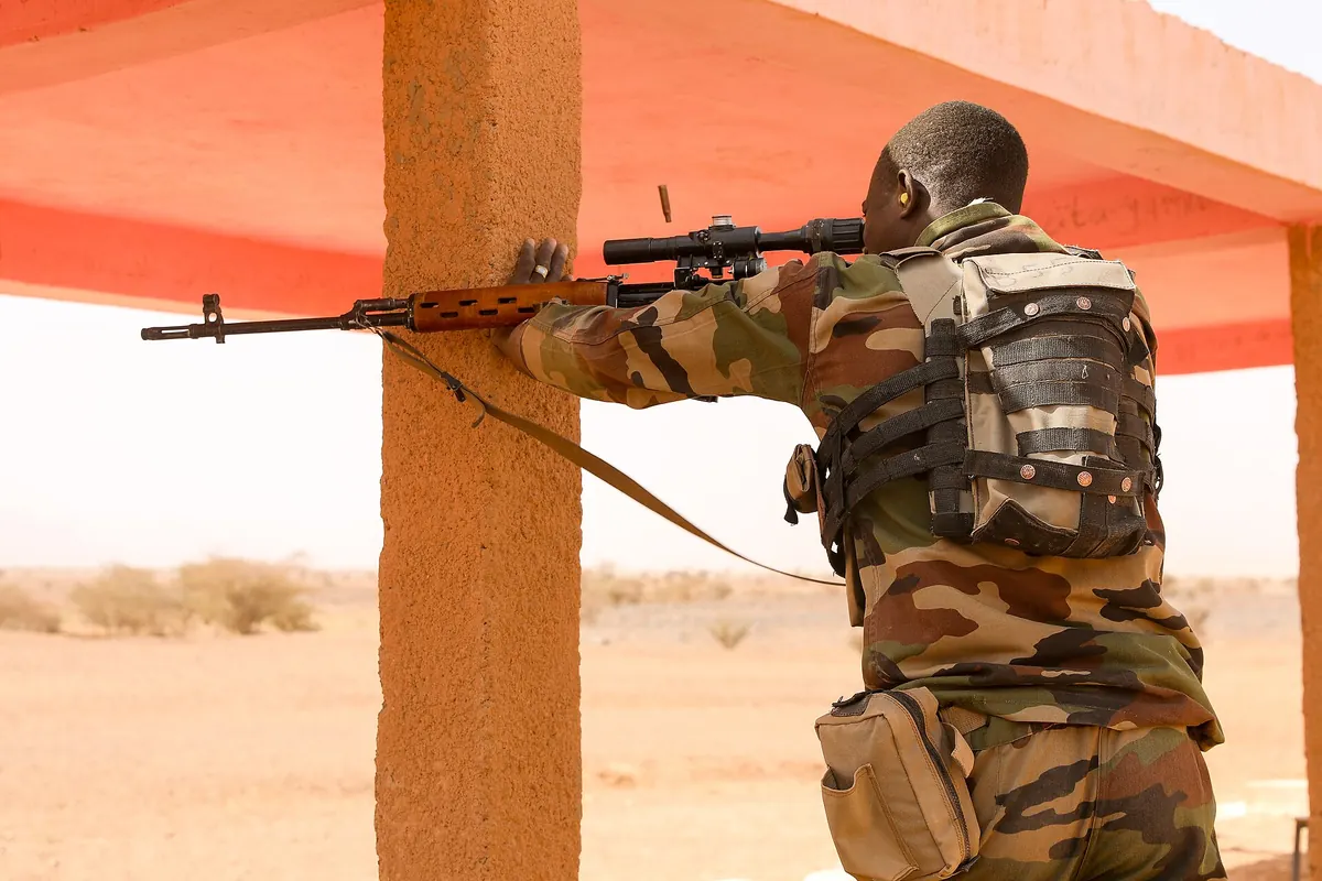 A soldier in camouflage aiming a rifle from behind a concrete pillar during training