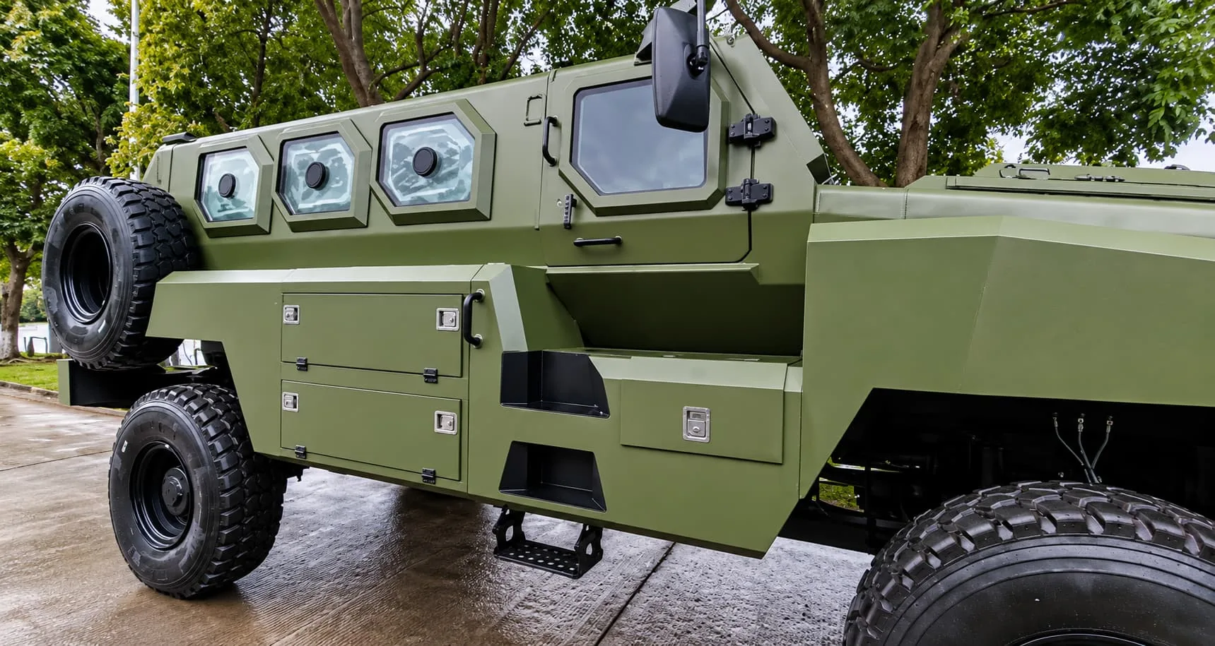Side view of an olive-green armoured personnel carrier with octagonal armoured windows, storage lockers and a spare wheel